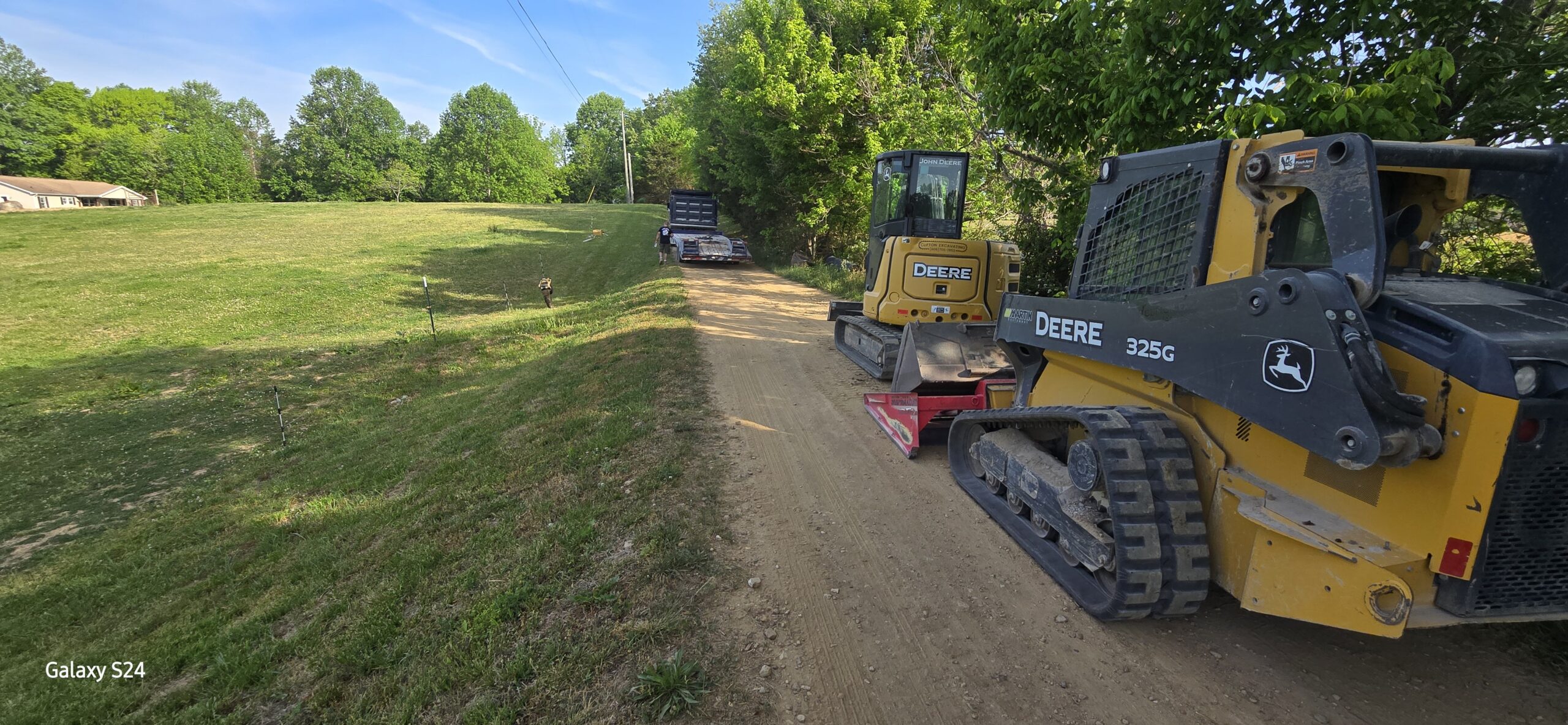 Two John Deere construction vehicles are parked on a dirt road beside a grassy field with trees and a house in the background under a clear, blue sky.