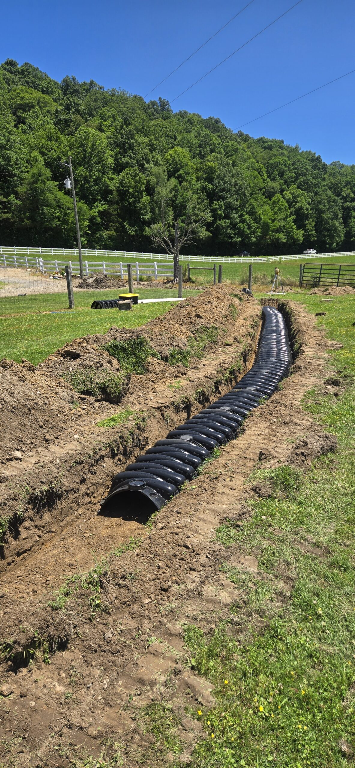 A black plastic drainage pipe is partially installed in a trench in a grassy field, with fencing and trees in the background under a clear blue sky.