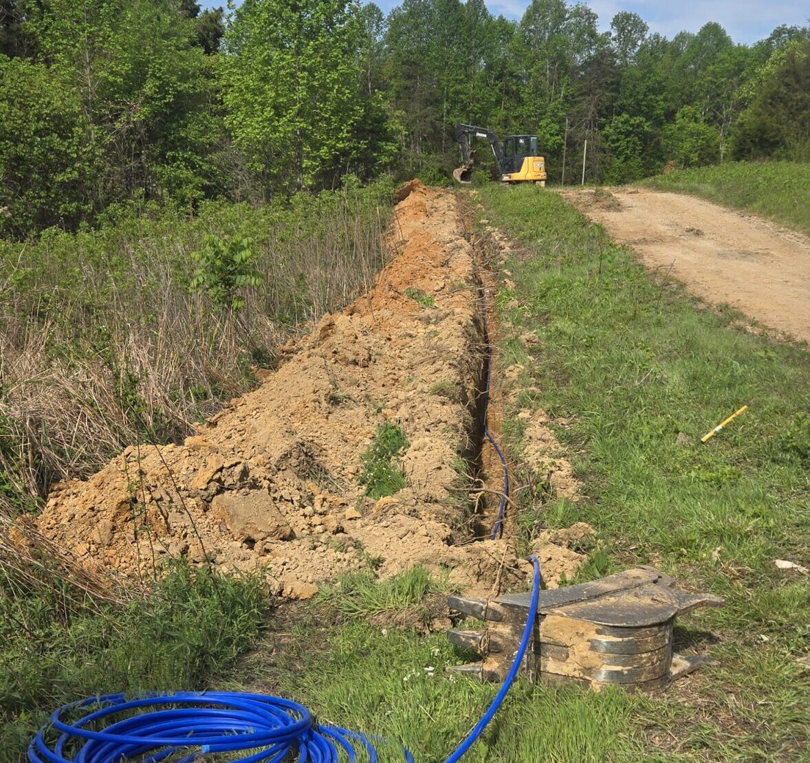 A trench runs alongside a dirt road with blue tubing coiled at the edge, surrounded by grass and trees under a partly cloudy sky.