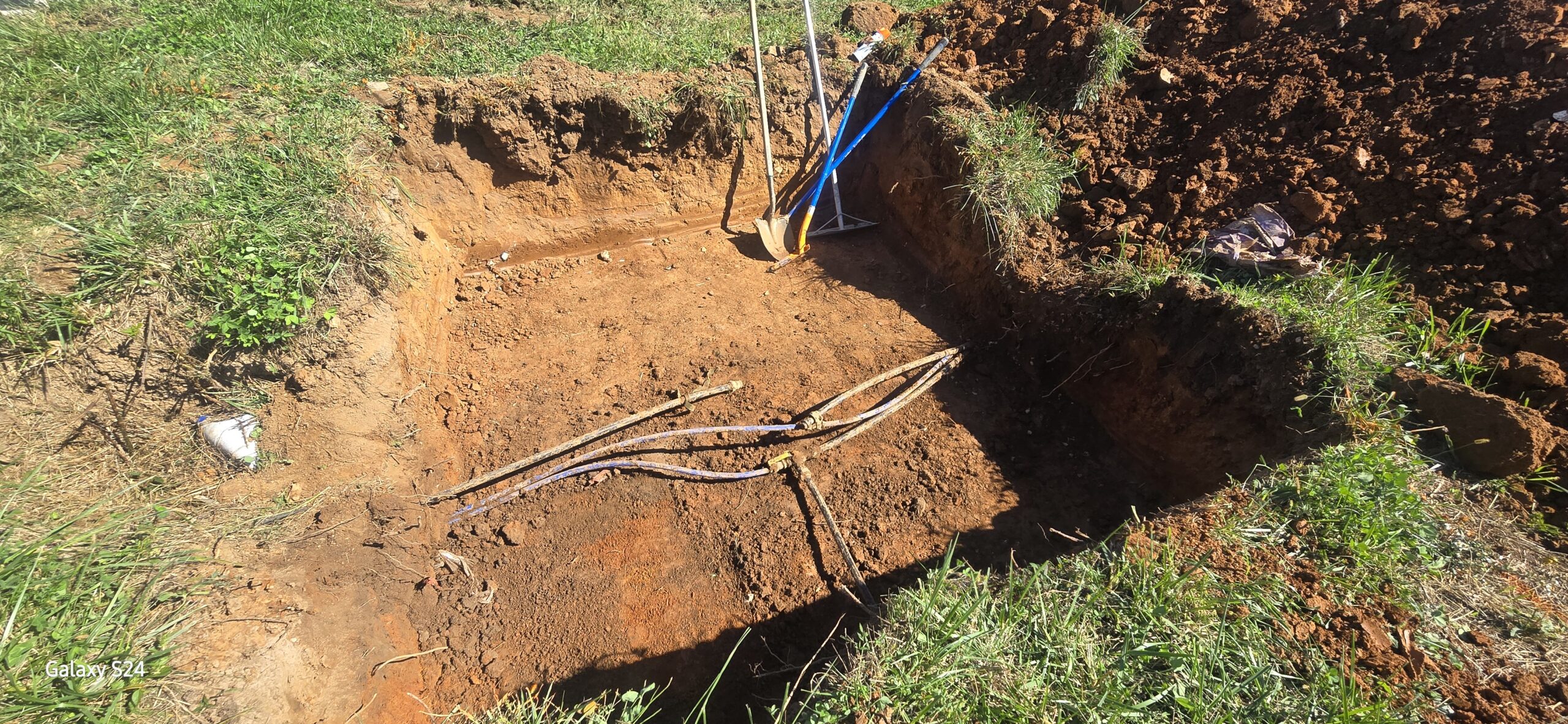 A rectangular hole dug in the ground shows several exposed pipes and some upright tools resting against one edge, surrounded by grass and soil.