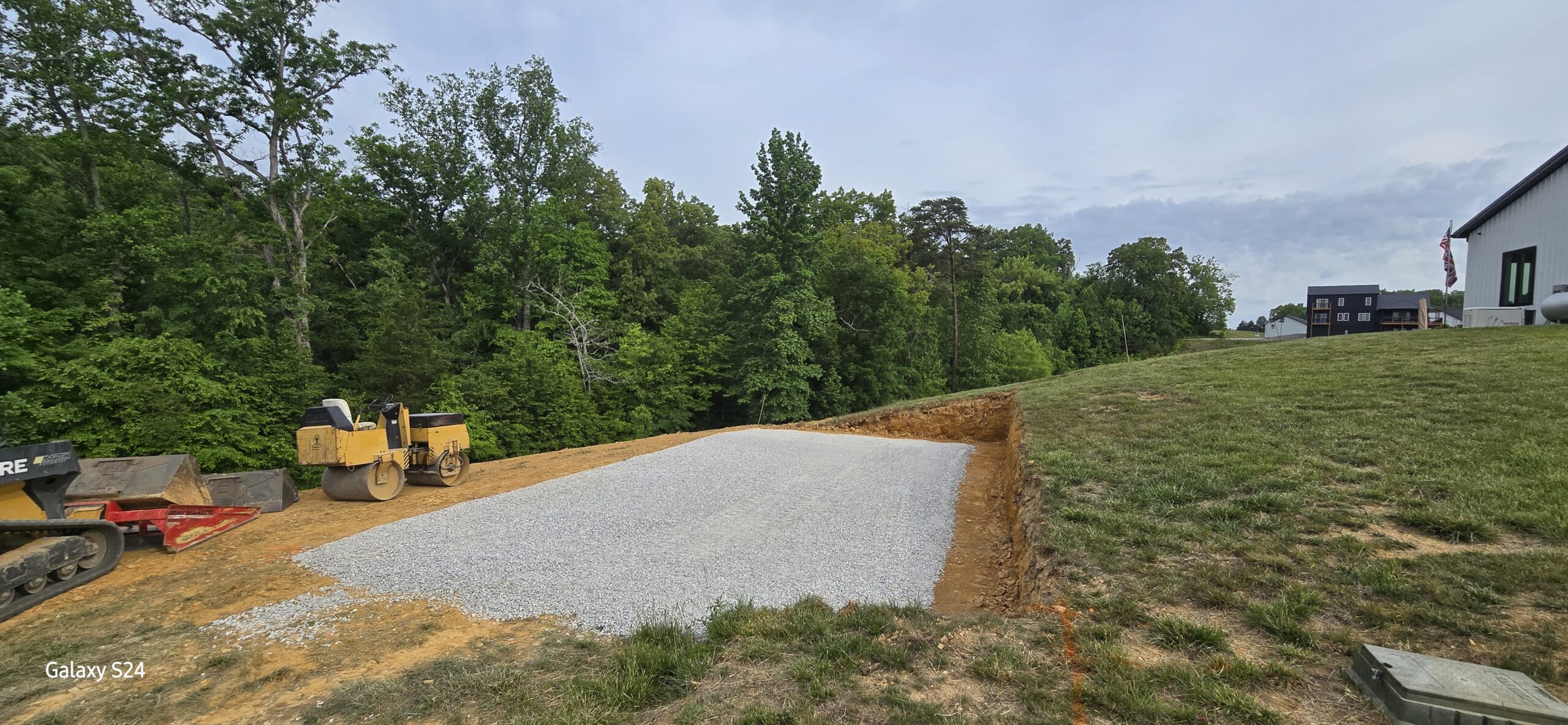 Construction site with compacted gravel foundation next to grassy slope, surrounded by trees and heavy machinery on the left side.