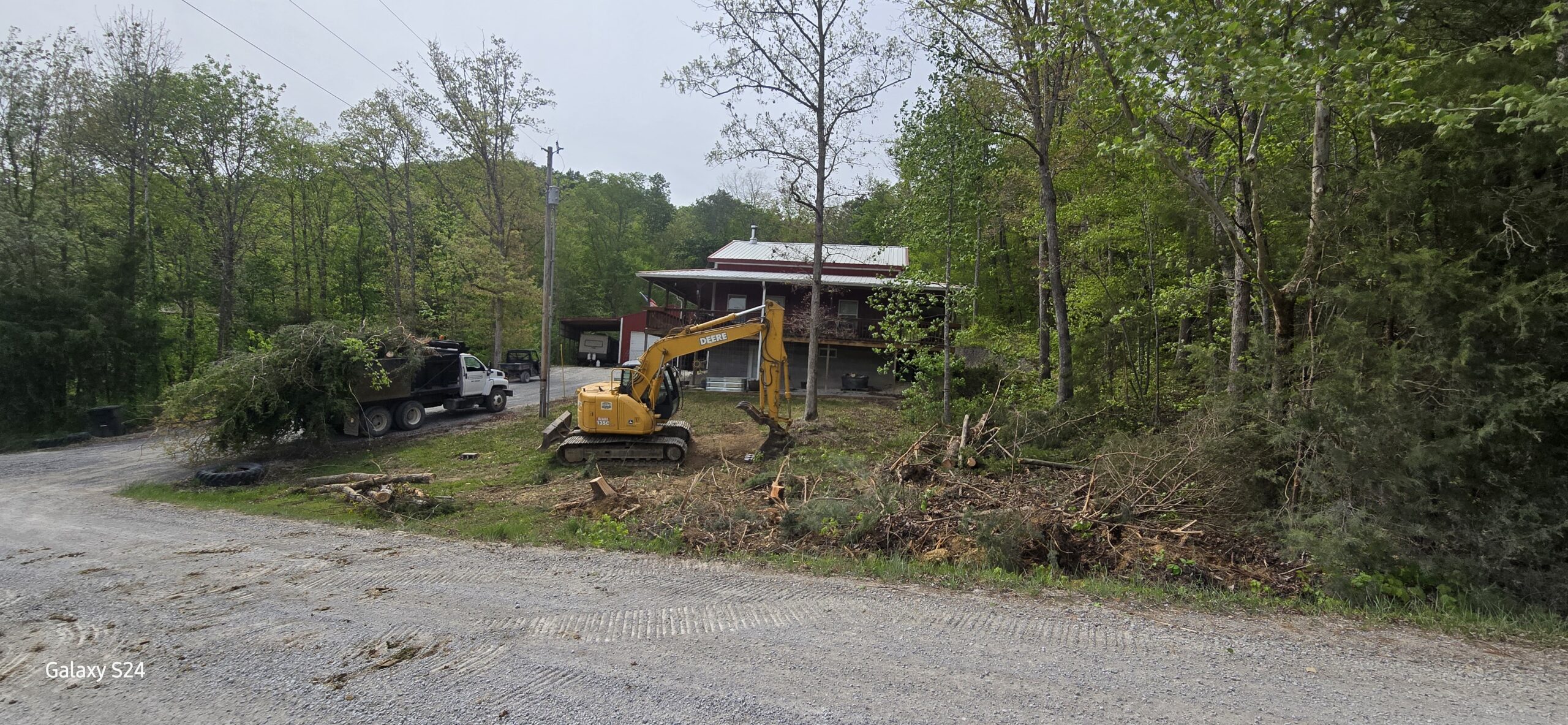 A yellow excavator clears brush near a gravel road, with a dump truck and a house in the background surrounded by trees.