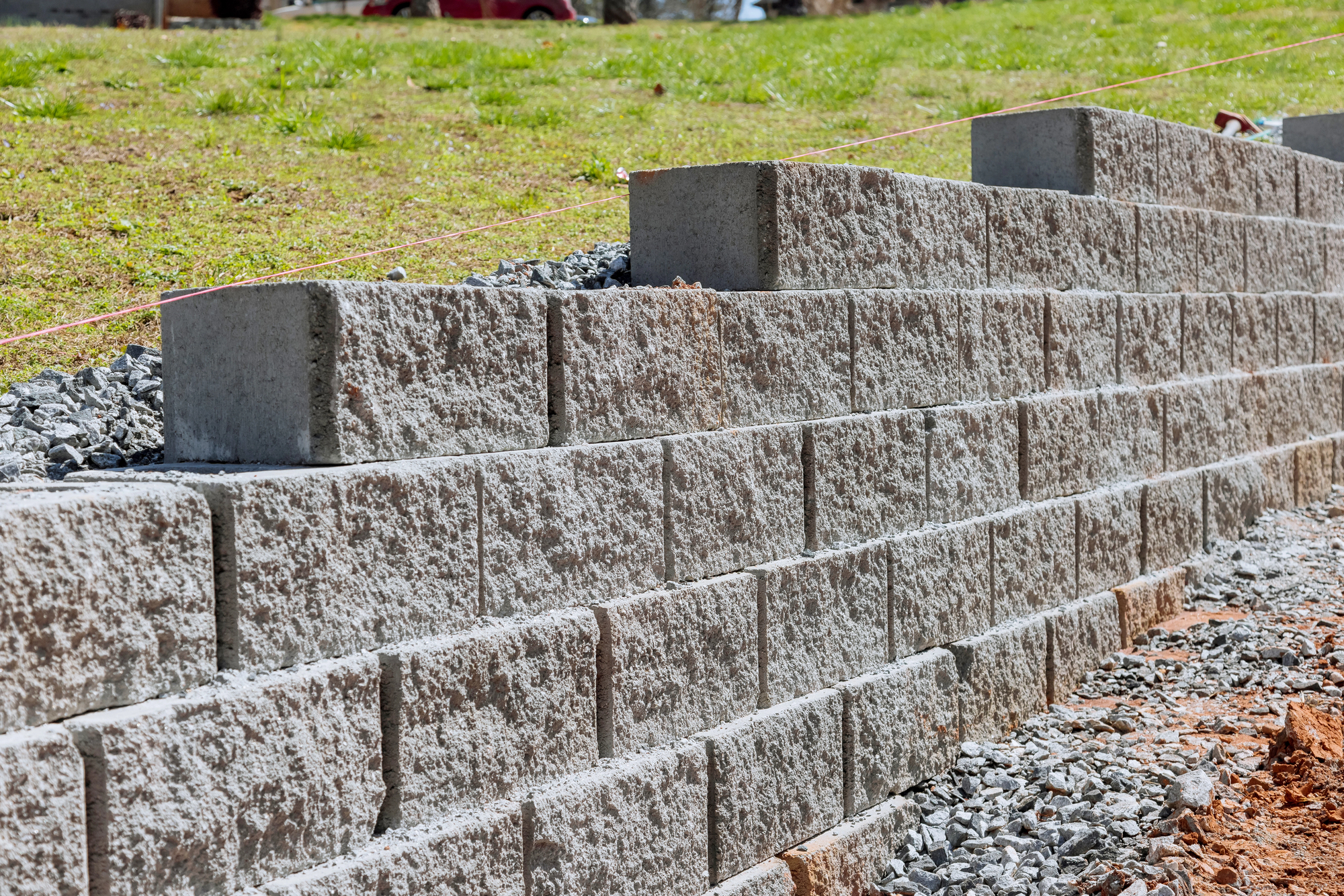 A close-up view of a retaining wall made of stacked concrete blocks with gravel and soil around the base on a sloped lawn.