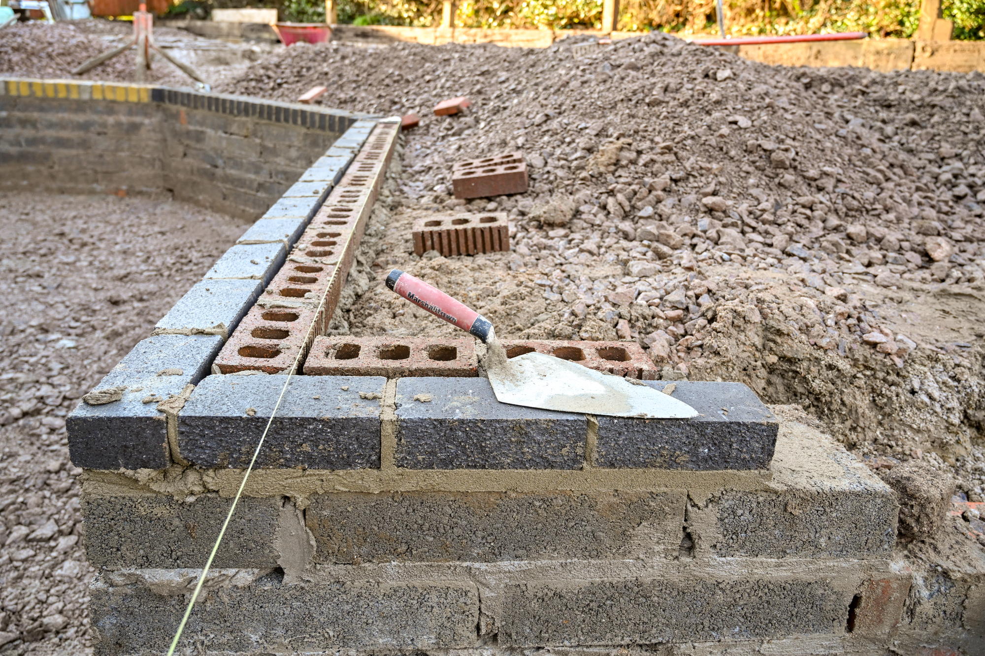A partially built brick wall at a construction site with a trowel resting on top and loose bricks and rubble in the background.