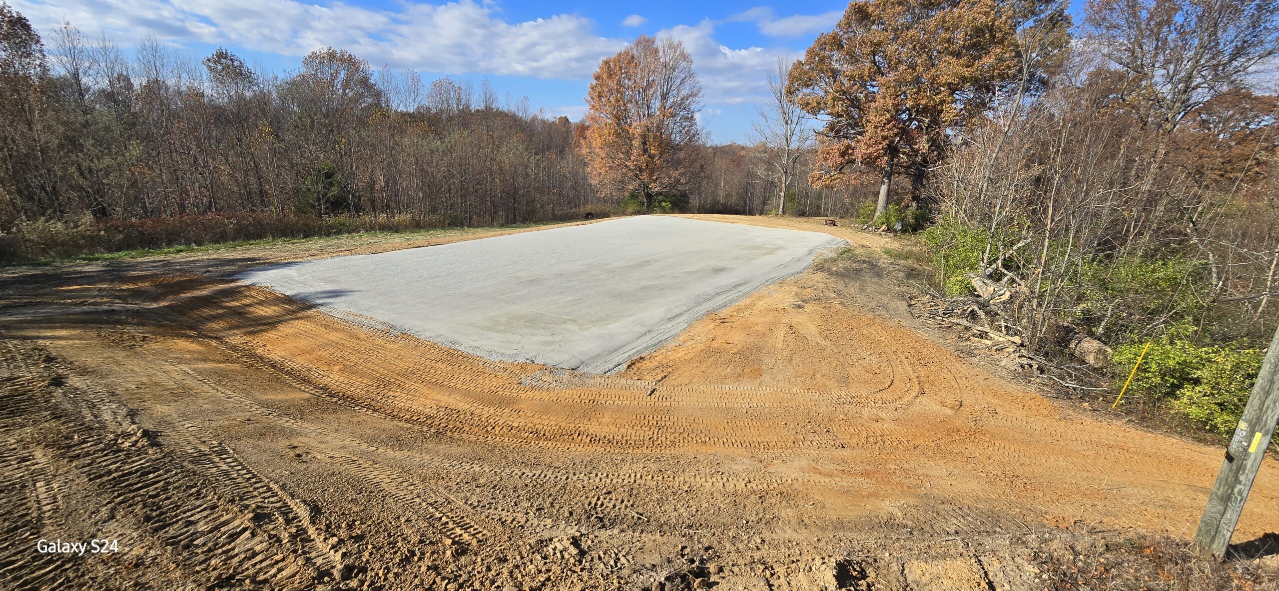 A freshly poured concrete slab sits in the middle of a cleared dirt area surrounded by trees with autumn foliage under a partly cloudy sky.